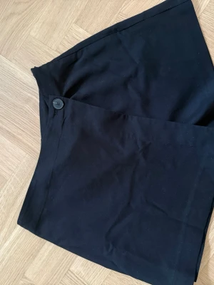 Svart skort med knappdetalj - Stilren svart skort med omlott-look och knappdetalj framtill. Klassisk rak passform och tillverkad i ett mjukt tyg som ger en snygg siluett. Perfekt för dig som vill ha shortskänsla men kjolens look.
