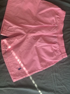 Rosa badbyxor med broderad logga - Säljer ett par ljusrosa badbyxor med elastisk midja och broderad logga i mörkblått framtill. Klassisk modell med fickor på sidorna och en bakficka. Perfekta för stranden eller poolen, tillverkade i snabbtorkande material.