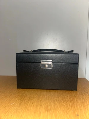 Smyckes skrin - Stilrent svart smyckeskrin i rektangulär form med handtag och lås framtill. Insidan är klädd i ljus textil och har flera fack för ringar, örhängen och andra smycken samt en liten spegel i locket. Perfekt för att organisera och förvara dina favoritsmycken.