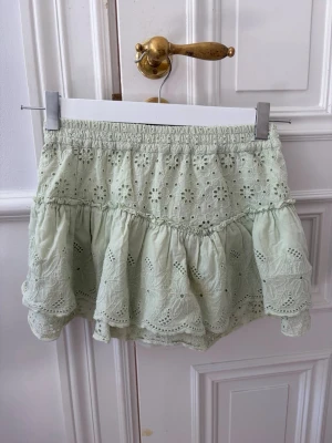 Ljusgrön volangkjol med broderi - Söt ljusgrön kortkjol med volanger och broderade blommönster. Kjolen har resår i midjan och är tillverkad i bomull, vilket gör den både luftig och bekväm. Perfekt för varma dagar och ger en romantisk känsla till din outfit.