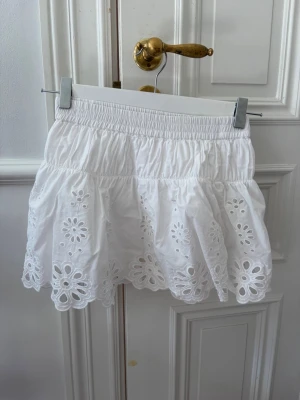Vit kortkjol med broderade blommor - Söt vit kortkjol med elastisk midja och broderade blommor längs kanten. Kjolen har volang och är gjord i bomull, perfekt för en fräsch och somrig look. Passar till både sneakers och sandaler.