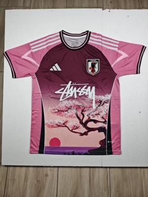 Japan Stussy rosa matchtröja med körsbärsträd - Unik rosa och vinröd matchtröja inspirerad av Japan, med körsbärsblom- och solnedgångsmotiv framtill. Tröjan har korta ärmar, rund hals och vita detaljer samt tryckt Stüssy-logga och Japan-emblem på bröstet. Tillverkad i ett lätt och snabbtorkande material.
