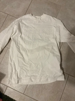 Vit långärmad topp Maison Margiela - Vit långärmad topp från Maison Margiela med ikoniskt siffror-tryck framtill och svart text på ryggen. Rund halsringning och relaxed passform. Perfekt för dig som gillar minimalistisk och stilren design.
