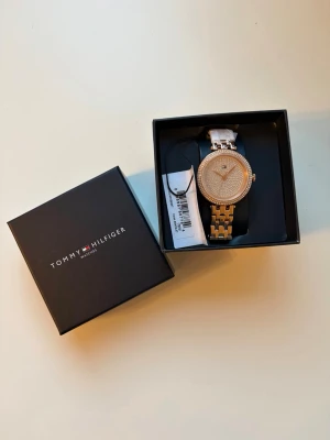 Guldig klocka Tommy Hilfiger - Snygg guldig klocka från Tommy Hilfiger med glittrig urtavla och stenar runt boetten. Länken är i metall och klockan har en elegant och modern design. Den är helt oanvänd och har till och med lappen kvar.