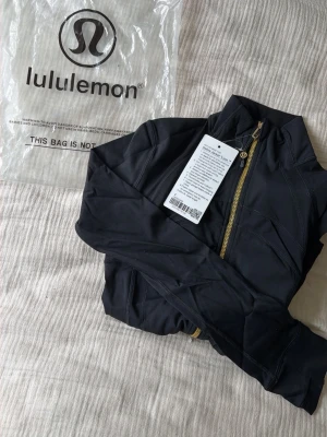 Svart träningsjacka från Lululemon stl 4 S  - Snygg svart träningsjacka från Lululemon med guldfärgad dragkedja och hög krage. Tillverkad i ett stretchigt funktionsmaterial som andas och sitter tight. Perfekt för gymmet eller löprundan. Stilsäker design med diskreta sömmar och logga på dragkedjan.
