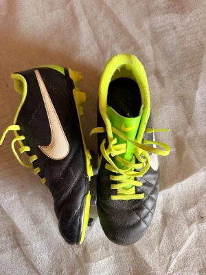 Nike svart/gul fotbollssko - Snygga fotbollsskor från Nike i svart med neon-gula detaljer och vita swoosh-loggor. Skorna har klassisk snörning, tålig ovandel i syntetmaterial och dobbar för spel på gräs. 