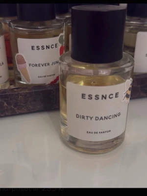 ESSNCE Dirty Dancing parfym - ESSNCE Dirty Dancing Eau de Parfum i en rund glasflaska med svart lock. Doften har en stilren vit etikett med svart text och en liten dekoration. Perfekt för dig som gillar unika och moderna parfymer.