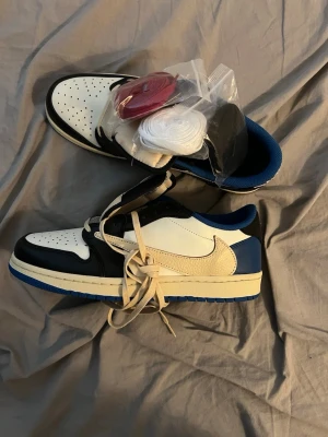 Travis Scott x Jordan 1 Low SP Fragments - Snygga sneakers i svart, vitt och blått med låg profil och kontrasterande detaljer. Skorna har en stor vit swoosh på sidan och kommer med extra skosnören i rött, vitt och svart. Ovandelen är i läder med perforerad tåbox och blå yttersula.