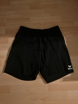 Svarta träningsshorts från Erima - Säljer ett par svarta träningsshorts från Erima med elastisk midja och logga på vänster ben. Perfekta för sport eller gym och tillverkade i ett lätt och bekvämt syntetmaterial.