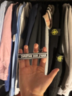 Christian Dior armband - Snyggt svart och vitt tygarmband från Christian Dior med texten 'CHRISTIAN DIOR J'ADIOR' broderad framtill. Armbandet har dekorativa tofsar i ändarna och justerbar knytning för perfekt passform. Perfekt accessoar för att lyfta din outfit.