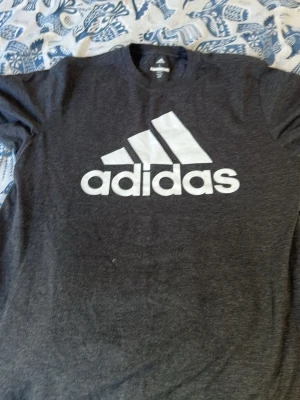 Mörkgrå Adidas t-shirt med logga - Snygg mörkgrå t-shirt från Adidas med stor vit logga på bröstet. Klassisk rund hals och korta ärmar. Perfekt för dig som gillar sportig stil och vill ha något bekvämt till vardags. Tillverkad i mjuk bomull.