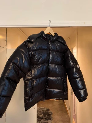 Svart pufferjacka från Moncler - Säljer en svart, glansig pufferjacka från Moncler med hög krage och dragkedja framtill. Jackan har två stora fickor med dragkedja och en Moncler-logga på ärmen. Perfekt för kalla vinterdagar och riktigt snygg streetstil.