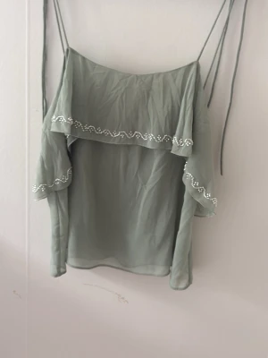 Ljusgrön blus med pärlor - Supersöt ljusgrön blus med axelband man justerar själv. Volangdetaljer med pärlor längs kanterna ger en extra touch. Tillverkad i ett tunt, luftigt material som känns perfekt för varmare dagar.