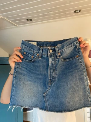 Blå jeanskjol från Levi's - Säljer en klassisk blå jeanskjol från Levi's med rå kant nertill och femficksmodell. Kjolen har bälteshällor, knappgylf och bakfickor med den ikoniska Levi's-detaljen. Perfekt för dig som gillar en avslappnad och trendig stil.