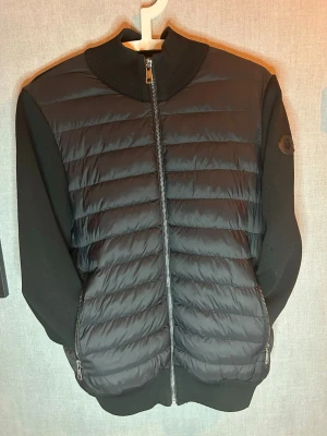 Black label moncler cardigan  - Snygg svart pufferjacka med quiltad front och rygg, ribbad krage och muddar. Jackan har dragkedja framtill och två fickor med dragkedja. Ärmarna är i ett mjukare tyg som kontrasterar mot den glansiga pufferdelen. Perfekt för kalla vinterdagar.