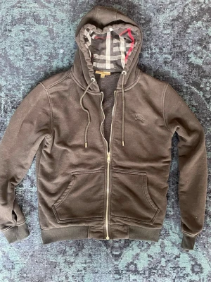 Svart hoodie med dragkedja från Burberry - Snygg svart hoodie från Burberry med dragkedja framtill och klassiskt rutigt foder i huvan. Storlek M men den passar bättre som S. Hoodien har fickor på magen, ribbade muddar och justerbar dragsko i huvan. Perfekt för en chill och stilren look. 
