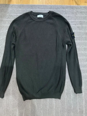 Mörkgrön stickad tröja Stone Island - Snygg mörkgrön stickad tröja från Stone Island med rund halsringning och ribbade muddar. Tröjan har lång ärm och den klassiska Stone Island-loggan på vänster ärm. Perfekt för dig som gillar stilrena och tidlösa plagg.