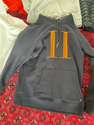 Marin blå hoodie från Les Deux - Snygg marinblå hoodie från Les Deux med stor orange romersk tvåa på bröstet. Klassisk känguruficka framtill och justerbar huva med snören. Tillverkad i mjukt bomullsmaterial som är skönt att ha på sig. Pris kan diskuteras 