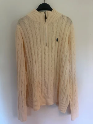 Beige kabelstickad tröja med halvzip - Kabelstickad tröja i beige med halvzip och hög krage. Tröjan har långa ärmar, ribbade muddar och ett diskret broderat logotyp på bröstet. Perfekt för lager på lager och riktigt snygg till jeans eller kjol.