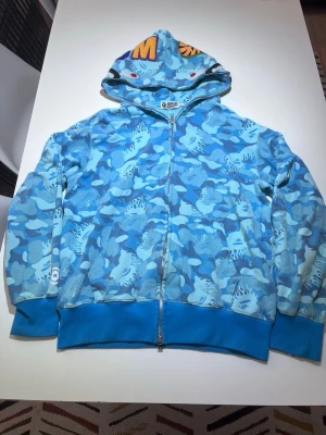 Blå camo hoodie från A Bathing Ape - Säljer en blå camo hoodie från A Bathing Ape med dragkedja och ikoniskt hajmotiv på huvan. Hoodien har broderade bokstäver och detaljer i flera färger på huvan samt ribbade muddar. Tillverkad i mjuk bomull, perfekt för streetwear-stilen.