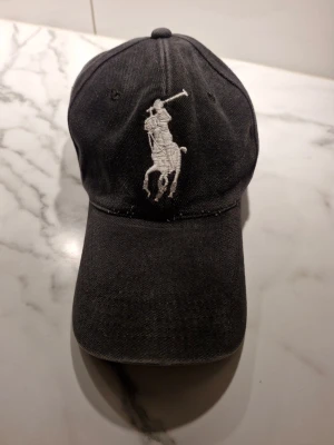 Svart keps från Polo Ralph Lauren - Snygg svart keps från Polo Ralph Lauren med den klassiska vita polospelaren broderad framtill och siffran 3 på sidan. Kepsen har böjd skärm och justerbar rem bak. Tillverkad i bomull för en bekväm och avslappnad stil.