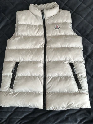 Vit dunväst från Moncler - Snygg vit dunväst från Moncler med klassisk quiltad design och hög krage. Västen har två svarta dragkedjefickor framtill och Monclers ikoniska logga på bröstet, Perfekt för lager-på-lager och ger en clean look till din outfit.
