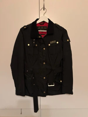 Svart Barbour jacka med bälte - Klassisk svart jacka från Barbour med guldiga knappar, bälte i midjan och flera fickor. Jackan har krage i manchester och detaljer som axelklaffar och Barbour-logga på bröstet. 