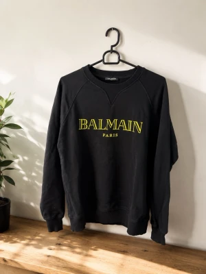 Svart sweatshirt från Balmain - Cool svart sweatshirt från Balmain med gul text på bröstet där det står 'BALMAIN PARIS'. Tröjan har rund halsringning, långa ärmar och ribbade muddar vid ärmslut och nederkant. Perfekt för en avslappnad och trendig look.