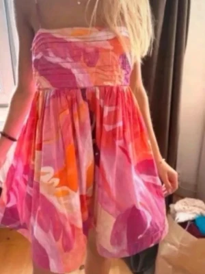 Ny trendig Färgglad kort sommarklänning från H&M  - Säljer en kort klänning med tunna axelband och färgstarkt abstrakt mönster i rosa, orange, lila och vitt. Klänningen har en luftig passform och är perfekt för varma dagar. Materialet känns lätt och svalt, troligen bomull eller polyester.