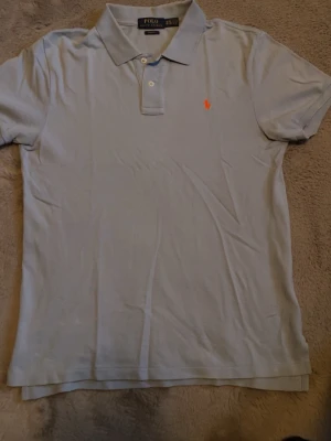 Ljusblå pikétröja från Polo Ralph Lauren - Klassisk ljusblå pikétröja från Polo Ralph Lauren med korta ärmar och krage. Tröjan har två knappar framtill och den ikoniska orange logotypen broderad på bröstet. Perfekt till jeans eller shorts för en clean look.