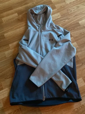 Grå och marinblå hoodie Under Armour - Snygg hoodie från Under Armour i grått och marinblått. Dragkedja framtill, huva med dragsko och två sidofickor. Logga på bröstet och svarta detaljer på ärmarna. Perfekt för chill eller träning. 