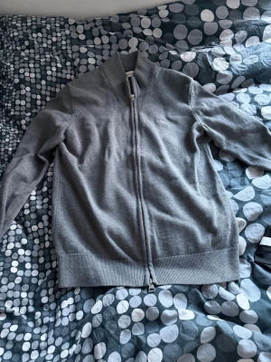 Grå ziptröja med hög krage - Snygg grå ziptröja med hög krage och ribbade muddar. Tröjan har hel dragkedja framtill och är tillverkad i ett mjukt, tunt stickat material. Perfekt att slänga på sig över en t-shirt för en clean look.