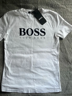 Vit t-shirt från Hugo Boss - Snygg vit t-shirt från Hugo Boss med stor svart BOSS-logga på bröstet. Klassisk passform och rund halsringning. Tillverkad i mjuk bomull som känns skön mot huden. Perfekt till jeans eller shorts för en clean och stilren look.