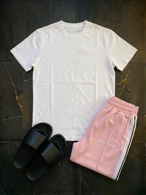 Rosa shorts från Palm Angels - Säljer ett par snygga rosa shorts från Palm Angels med vit sidrand och elastisk midja. Shortsen har en avslappnad passform och är perfekta för sommardagar. Ikonisk logga broderad på sidan för extra streetwear-vibe. Shortsen är använda men har inga tydliga tecken på användning. Perfekt för dig som vill se stilren ut i sommar.
