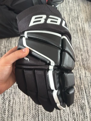 Svarta hockeyhandskar från Bauer - Säljer ett par svarta hockeyhandskar från Bauer med vita detaljer och kraftig vaddering. Handskarna har en modern design och är tillverkade i slitstarkt syntetmaterial, perfekta för ishockeyträning och match. Och aldrig använda 