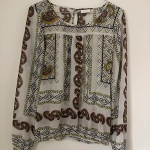 Genomskinlig blus i strlek M ifrån VILA - En något genomskinlig blus ifrån VILA i storlek M. Vit i grunden med mönster i olika färger. Den har ett resårband längst ner och tröjan knäpps vid handlederna och i nacken med silverknappar. 