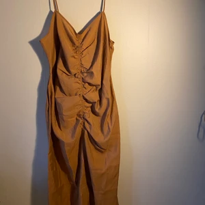 Brun knä lång klänning  - En V-Ringad klänning men regler bara smala axelband och rynkad söm på fram och baksidan och en dold dragkedja på sidan. Klänningen är aldrig använd, den är i storlek 50 men liten i storleken. 