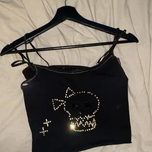 Svart croppad top med dödskalle - Svart croppad topp med dödskalle i rhinestones på och vita sömmar runtom (y2k likt), ett hål vid höger axelband💀☠️✨💗