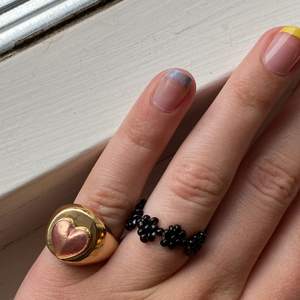 Svart handgjord ring av elastisk tråd, med gulligt blommönster! 🤍 bredvid syns vår Heart Ring som kostar 99kr