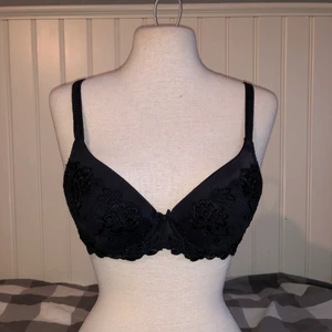 Svart bh. Storlek står ej, men skulle säga 75B - Söt svart bh med blommönster av spets och en liten rosett mellan kuporna. Märket är Triumph. Säljer för att den är för liten! Lite vadderad.