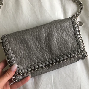 Axelremsväska från Tiamo - Snygg grå axelremsväska i skinnimitation kantad med silverfärgad kedja. Ett fack med dragkedja på insidan. Den avtagbara axellänken gör att väskan även kan bäras som en clutch. Axellänk 105 cm. Längd 21 cm, höjd 14 cm, bredd 5cm. Väskan är i bra skick😊
