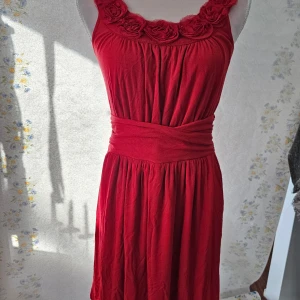 Röd klänning från Sara Kelly - En elegant röd klänning från Sara Kelly, designad av Ellos. Klänningen har en vacker detalj med rosor runt halsringningen och en bred midjeband som framhäver figuren. Den är ärmlös och har en mjuk, flödande passform.