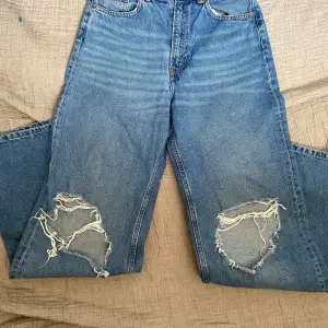 Ett par blå jeansbyxor med slitningar på båda benen. Byxorna har en hög midja och en lös passform.