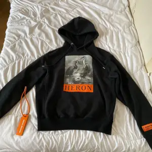 En svart hoodie från Heron Preston med en tryckt bild av två hägrar och texten 'HERON' i orange på framsidan. Hoodien har en justerbar huva och ribbade muddar vid ärmslut och nederkant. Märkesetikett i orange på ärmen.