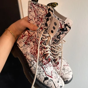 Dr. Martens skor - Dr Martens skor med ”färgstänk”. Skorna är vita i grunden och har svart och rött ”färgstänk. Använda fåtal gånger men har fått en slitning på vänstra skon (se bild) utöver de är dom som nya.