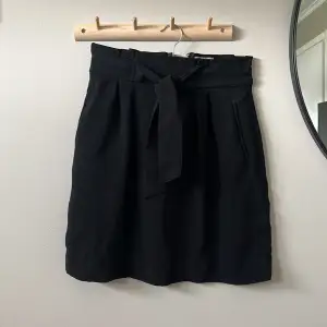 Kort svart kjol med knytskärp. Stäng med dragkedja på baksidan. Fickor på båda sidor
