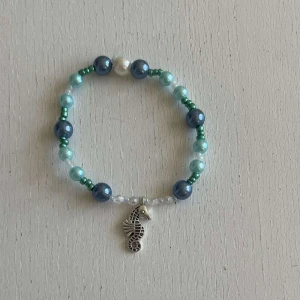 Egengjort pärlarmband  - Ett egengjort pärlarmband med färgerna blå, vit och grön i olika nyanser med en blandning pärlsorter. Armbandet har en berlock i silver som avbildar en sjöhäst.  