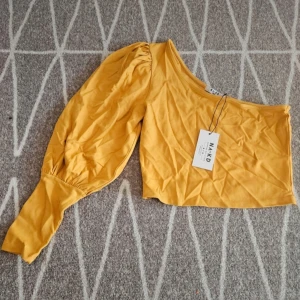 Ny croppad one shoulder topp  - Jättefin croppad one shoulder topp med puffärm. Färgen är senapsgul. Aldrig använd men lite skrynklig då den legat i lådan. Kommer från ett djur och rökfritt hem.