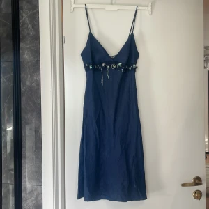 Mörkblå klänning från Blu Sand - En elegant mörkblå klänning från Blu Sand, tillverkad i Italien. Klänningen har smala axelband och en dekorativ blommig detalj under bysten. Den är ärmlös och har en kort längd, materialet är linne vilket gör den perfekt för festliga tillfällen och sommardagar. storleken 42 motsvarar s/m i storlek 