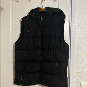 Svart väst med huva  - Svart väst med huva. Dragkedja med fickor vid sidorna och snören längs huvan. Puffer väst. Använts Max 5 gånger. Inga synliga fläckar eller märken. Pris kan diskuteras. 